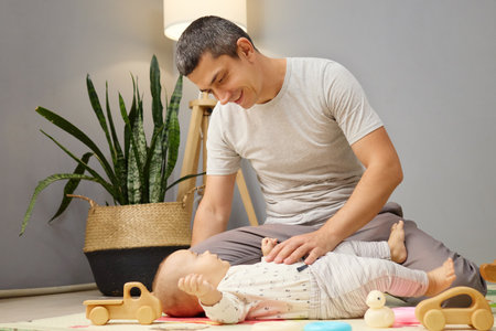 Young father holding his newborn child playing with kid while sitting on floor in living room man wearing casual clothing enjoying time together at home stroking the baby's tummyの写真素材
