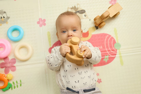 Top view of funny baby wearing bodysuit lying on carpet inside nursery biting toy while teething feeling pain in his mouth playing with eco-friendly toy carの写真素材