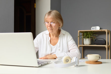 Mature woman with glasses focused on laptop typing at home office displaying serious concentration working online using modern technology in quiet interior settingの写真素材