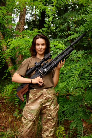 A woman stands in a forest, confidently holding a rifle. She wears a military-style outfit and shows a calm demeanor amidst the vibrant green foliage surrounding her.の写真素材