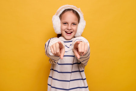 Cheerful little girl with fur headphones pointing finger choosing best choice looking at camera with...