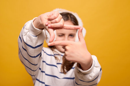Little girl wearing fur headphones framing her view with fingers raised focused on capturing moment with creativity and imagination isolated on yellow backgroundの写真素材