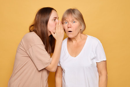 A younger woman leans in close, whispering to an older woman who looks shocked and intrigued, both against a vibrant yellow background conveying a sense of intrigue and surprise.の写真素材