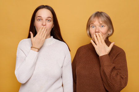 Two women stand side by side, surprised looks on their faces as they cover their mouths with their hands, set against a cheerful yellow backdrop that adds warmth to the moment.の写真素材