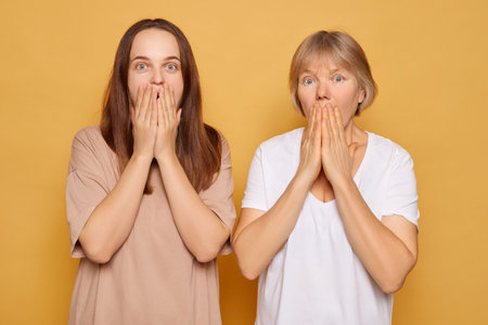 The two women display surprised expressions with their hands covering their mouths, set against a bright yellow backdrop, capturing a lighthearted and spontaneous moment.の写真素材