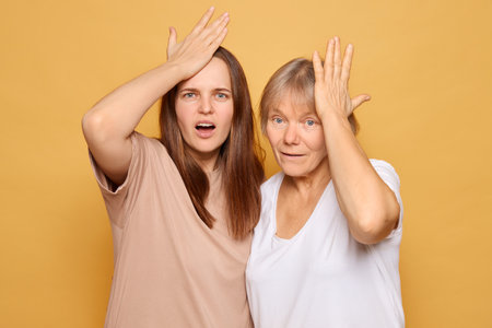 Two women of different ages express surprise by touching their foreheads, looking astonished against a bright yellow backdrop, creating a fun and engaging atmosphere.の写真素材