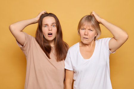 Two women display expressions of confusion and surprise, standing side by side with their hands on their heads, set against a solid yellow background that emphasizes their emotions.の写真素材