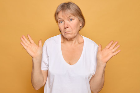 A woman wearing a simple white shirt displays a confused expression while raising her hands in a shrugging gesture, set against a vibrant yellow background.の写真素材