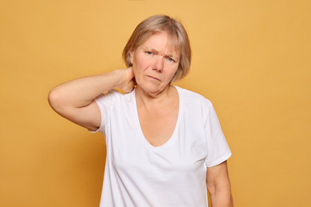A mature woman stands with a pained expression, gripping her neck, suggesting discomfort or tension, set against a vibrant yellow backdrop that enhances the mood.の写真素材