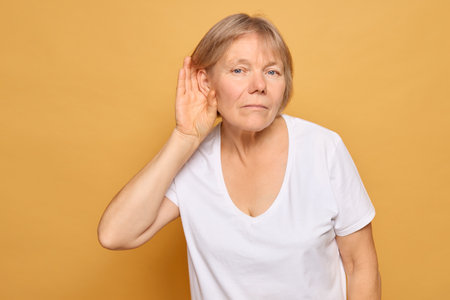 A woman with short blonde hair is actively listening, her hand cupped to her ear, indicating curiosity or interest in what someone is saying against a vibrant yellow backdrop.の写真素材