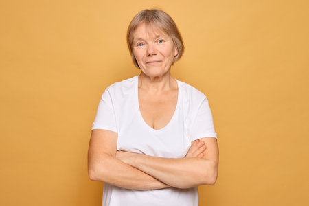 A woman with short hair smiles gently while standing with her arms crossed. She wears a plain white t-shirt and is set against a bright yellow backdrop, radiating warmth and confidence.の写真素材