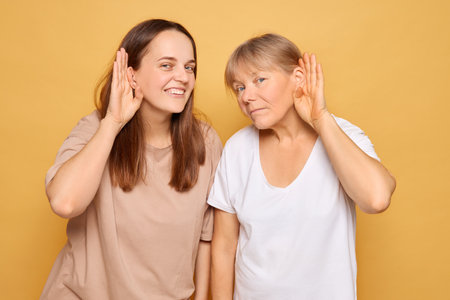 Two women pose together against a vibrant yellow backdrop, each playfully cupping their ears while leaning forward, suggesting a humorous attempt to hear something better.の写真素材