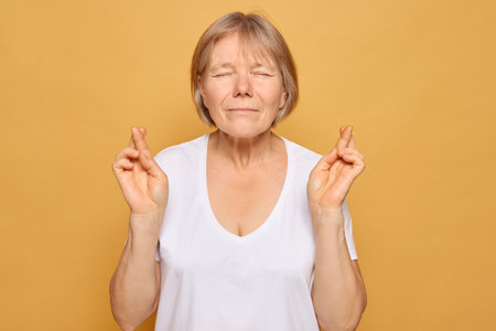 A smiling woman with closed eyes gently crosses her fingers, conveying a sense of hope and good fortune against a warm yellow backdrop that radiates positivity.の写真素材