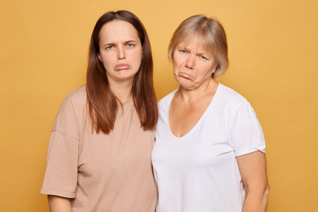 In a brightly lit space, two women stand together, both displaying serious and neutral expressions. They wear casual clothing and appear to be sharing a moment of contemplation.の写真素材