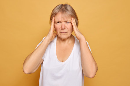 A middle-aged woman with short hair expresses discomfort and concern, holding her head with both hands. She stands against a yellow backdrop, conveying a sense of stress.の写真素材