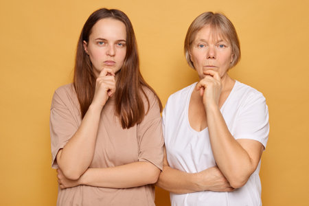 Two women stand side by side with serious expressions, their fingers on their lips, suggesting deep thought or pondering, set against a bright yellow background.の写真素材