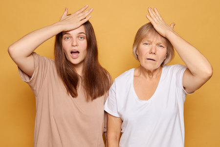 Two women stand side by side, one younger and one older, each with a hand on their forehead, displaying expressions of surprise and confusion while against a cheerful yellow backdrop.の写真素材