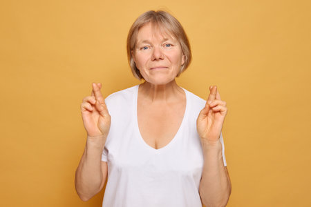 In front of a vivid yellow backdrop, a woman smiles gently, crossing her fingers, symbolizing hope and good luck, radiating a warm and inviting energy.の写真素材