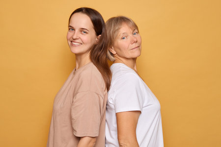 Two women stand back to back, one smiling brightly while the other gazes thoughtfully. Their contrasting expressions add depth to the warm yellow backdrop.の写真素材