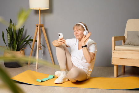A mature woman sits comfortably on a yoga mat at home, interacting with her smartphone while wearing headphones and participating in a virtual fitness session, surrounded by plants.の写真素材