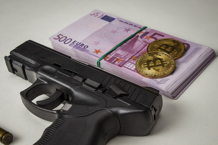 Black pistol with a pack of euros and coins in the form of bitcoin on a white tableの写真素材