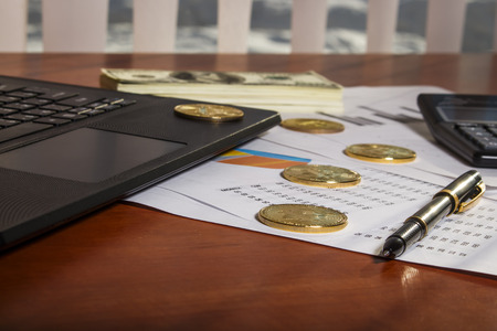 Laptop stands on the table near a pack of dollars and bitcoins in the officeの写真素材