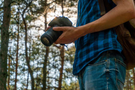 Young man in a blue plaid shirt with a camera in his hands in the coniferous forest