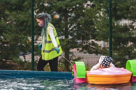 Petrozavodsk, Russia - 1 August 2020. a young woman an amusement Park worker in a protective mask pulls out a stuck boat with a childのeditorial素材