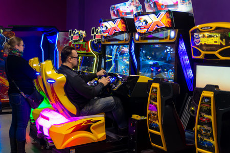 Saint-Petersburg, Russia - 5 January 2024. A man is playing a racing game in a brightly lit room. A woman is standing nearby. The room is filled with various video game machines, including a racing game with a yellow and black theme