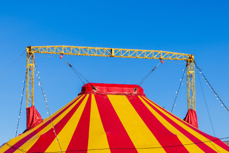 Red and yellow striped circus tent top held by yellow steel rigging against a clear blue sky.