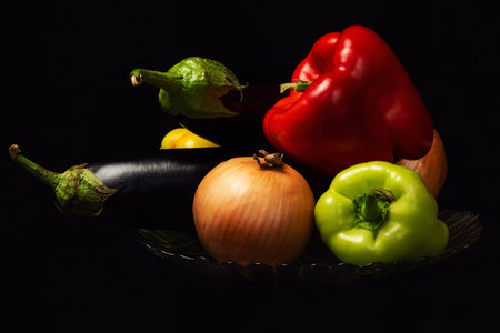 Fresh organic eggplants, tomatoes, peppers and onions on a glass plate with a black backgroundの写真素材