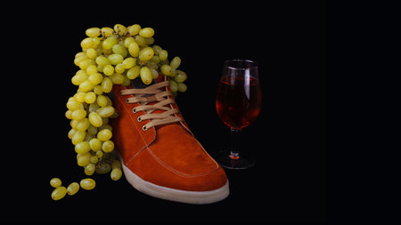 A bunch of white ripe grapes hangs from a man's suede autumn boot and a glass of wine. Studio art still life with the combination of clothes and food.の写真素材