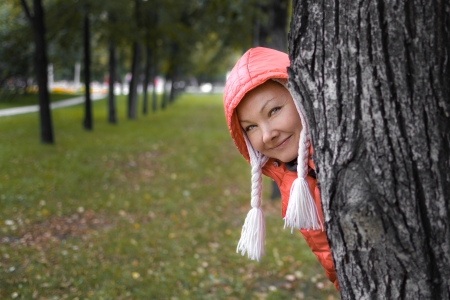 Young woman in red jackey looks out of the autumn park treeの写真素材