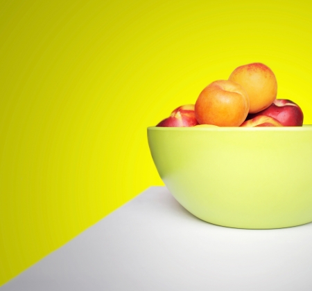 Fresh nectarines and apricots inside a green modern bowl on the table on yellow backgroundの写真素材