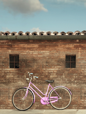 Bicycle parked and leaning on an old brick wall with two small windows, the roof and the skyの写真素材