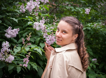 Young girl is standing near a blooming lilac bush and looking at camera.