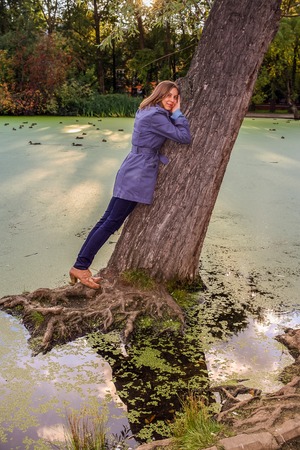 Young girl is standing leaning against a large bending down tree in water , and looking at camera in autumn evening.の写真素材