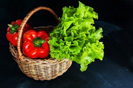 Ripe red peppers and bunch of green salad in wicker basket on dark background.の写真素材