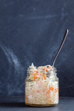 One glass jar with sauerkraut and metallic spoon is standing on dark table on dark background.の写真素材