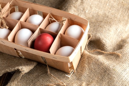Open eco-friendly wooden box with white eggs and one red Easter egg on rough burlap.