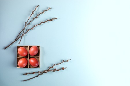 Red Easter eggs in cardboard box with willow branches on pale blue background.