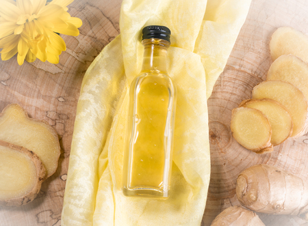 Closed glass bottle of ginger oil on yellow cloth in center of wooden saw cut with ingredients.
