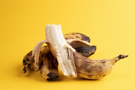 Close-up one peeled banana and two unpeeled overripe blackened ugly bananas on yellow background.