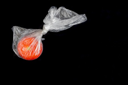 One red tomato in transparent closed plastic bag on black background.