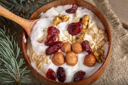 Close-up wooden bowl with yogurt with rolled oats, nuts and dried cranberries on burlap.