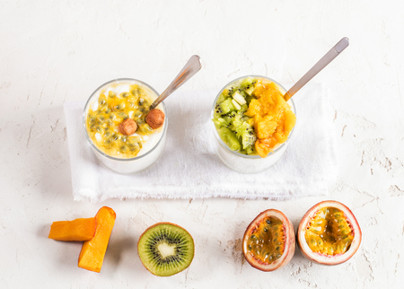 Two glasses with white yogurt and fruit puree of passion fruit, kiwi, mango and with metallic spoons and half fruits on white textured background. Top view.の写真素材