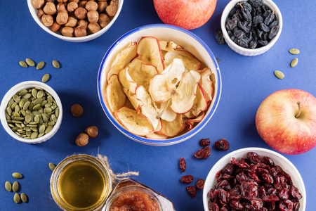 Ceramic cup with apple healthy homemade chips surrounded by white cups with healthy snacks, fresh apples and jars of jam and honey on bright blue background. Natural food. Fruit healthy snack. Top view, flat lay.の写真素材