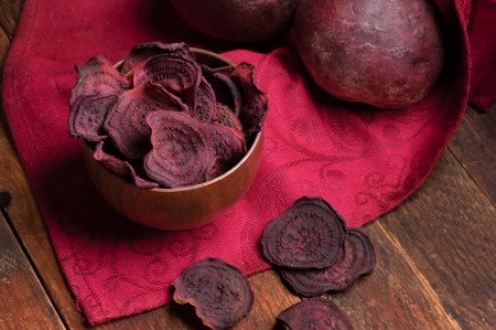 Beetroot chips in wooden bowl and whole beetroots on red napkin on old wooden table.