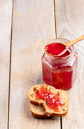 Toasts with homemade strawberry jam, open glass jar and wooden spoon full of jam on wooden table with copy space. Natural homemade dainty.の写真素材