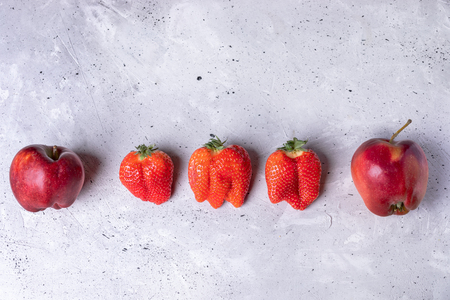 Ugly two apples and three strawberries are lying in row on grey concrete background.の写真素材